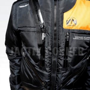 May include: Black jacket with a zipper closure, multiple pockets, and a yellow and orange accent on the upper right chest. The jacket has the text "DRAWERIDGE" and "SHIPBOARD USE APPROVAL SURVEILLANCE MARKING" printed on it. The jacket has a black strap with a silver zipper.