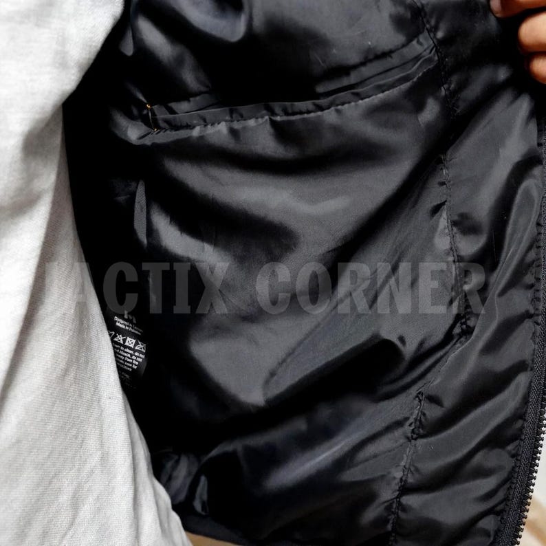 May include: Close-up of the inside of a black jacket. The interior lining is smooth and shiny, with a small pocket visible. The jacket is partially unzipped, revealing a gray shirt underneath. The text "CTIX CORNER" is visible.