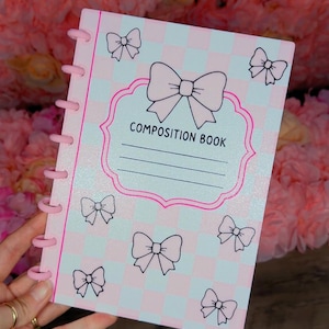 Puede incluir: Un cuaderno de composición a cuadros rosas y blancos con una serie de lazos de contorno negro esparcidos por la portada. El centro muestra una etiqueta decorativa con el texto COMPOSITION BOOK y tres líneas horizontales para escribir.