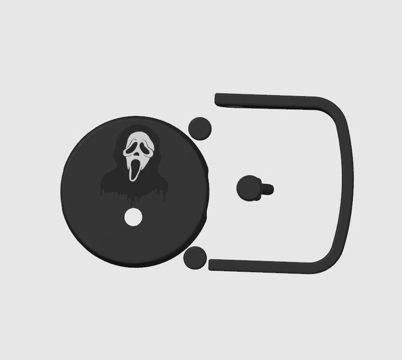 May include: A black belt buckle featuring a circular design with the iconic Ghostface mask from the Scream horror film series. The buckle has a rectangular frame and a pin for closure.