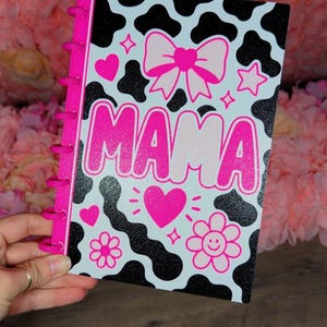 May include: A spiral-bound notebook featuring a black and white cow print pattern with glittery accents. The cover displays the word MAMA in large pink letters, surrounded by pink graphic elements including a bow, hearts, stars, and a smiley flower.