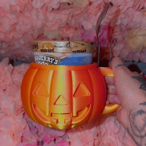 Puede incluir: Una taza de color naranja brillante con forma de calabaza y una cara de linterna tallada, que contiene un bote de helado Ben & Jerry's. Una cuchara de metal plateado está insertada en el lateral de la taza, que tiene un asa y acabado brillante.