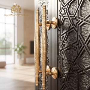May include: Close-up of a black door with ornate gold-colored handles. The handles are long and cylindrical with detailed engravings. The door features a geometric pattern. The logo "HANDMADEBYBRASS" is visible in the upper left corner.