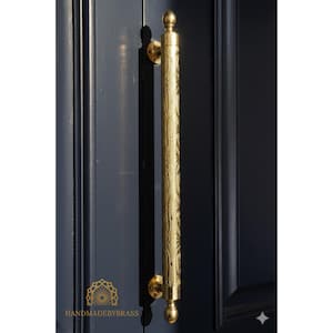 May include: A gold-coloured door pull with an ornate, textured design. The long, cylindrical handle is mounted on a dark blue door. The pull has a rounded top and bottom, and is attached to the door with circular mounts. The text "HANDMADEBYBRASS" is visible.