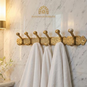 May include: A gold-colored, ornate wall-mounted hook rack with six hooks, holding white towels. The rack has decorative floral patterns and the text "HANDMADEBYBRASS". The hooks are mounted on a marble wall.