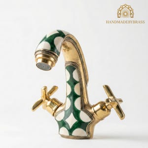 May include: A brass tap with a decorative design. The tap features a curved spout and cross-shaped handles. The design includes green and white diamond-shaped patterns. The tap has a gold finish and is set against a white background.