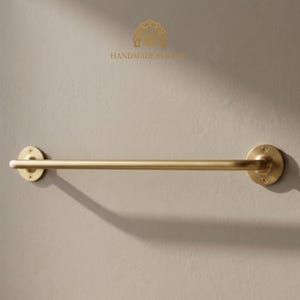 K&ouml;nnte beinhalten: Ein goldfarbener Handtuchhalter an einer hellbeige Wand. Die Stange ist zylindrisch mit kreisf&ouml;rmigen Montageplatten an jedem Ende. Der Text "HANDMADEBYSIRES" befindet sich &uuml;ber der Stange.