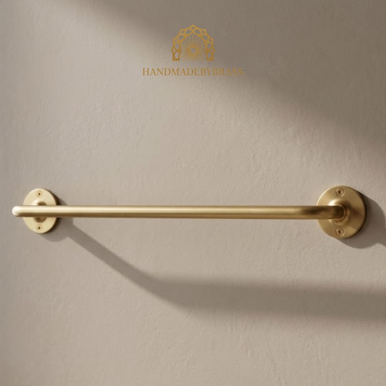 K&ouml;nnte beinhalten: Ein goldfarbener Handtuchhalter an einer hellbeige Wand. Die Stange ist zylindrisch mit kreisf&ouml;rmigen Montageplatten an jedem Ende. Der Text "HANDMADEBYSIRES" befindet sich &uuml;ber der Stange.
