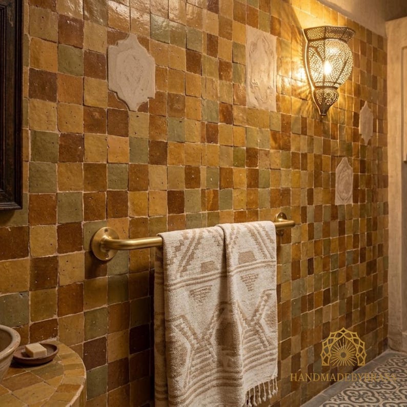 K&ouml;nnte beinhalten: Eine Badezimmerwand mit quadratischen Fliesen in Braun-, Gr&uuml;n- und Gelbt&ouml;nen. Ein Handtuchhalter aus Messing h&auml;lt ein beiges Handtuch mit geometrischem Muster. Eine dekorative Wandleuchte mit Muster ist an der Wand angebracht.