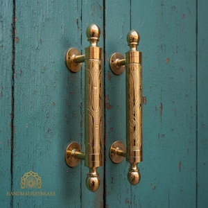May include: Two brass door handles with intricate engraved designs. The handles are cylindrical with decorative finials and circular mounting plates. The background is a weathered teal-colored wooden door. The text "HANDMADEBYBRASS" is visible.