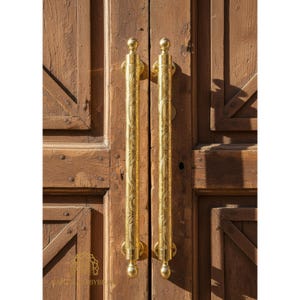 Könnte beinhalten: Nahaufnahme einer Holztür mit zwei verzierten, goldfarbenen Türgriffen. Die Griffe sind lange, vertikale Stangen mit dekorativen Mustern und abgerundeten Enden. Die Tür besteht aus gealtertem, braunem Holz mit sichtbarer Maserung.