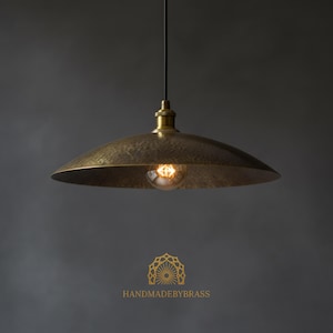 May include: A brass pendant light fixture with a hammered texture. The light has a wide, shallow shade and a visible Edison-style bulb. The fixture hangs from a black cord. The text "HANDMADEBYBRASS" is at the bottom.