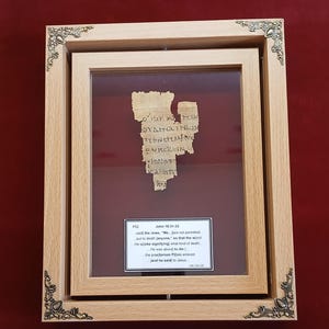 May include: A framed ancient papyrus fragment with Greek text, presented in a wooden frame with decorative corner details. A label below the papyrus provides context, including the text "John 18:31-33" and the date "100-150 CE".
