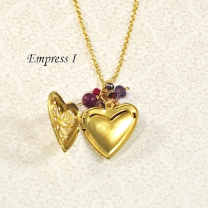 May include: Gold heart-shaped locket necklace with a filigree design on the inside of the locket. The locket is adorned with small purple and red beads.