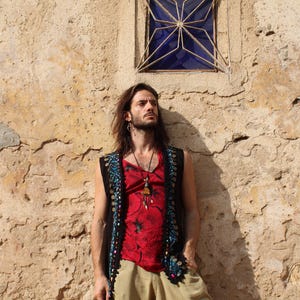 Puede incluir: Una persona con una camiseta roja con estampado floral, un chaleco negro con adornos azules y dorados y pantalones color beige. La persona está de pie frente a una pared texturizada de color marrón claro.
