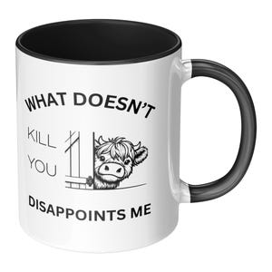 May include: White ceramic coffee mug with a black rim and handle. The mug has a black and white illustration of a cow peeking over a fence with the text "What doesn't kill you disappoints me."
