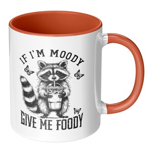 May include: White ceramic mug with a burnt orange interior and handle. The mug features a black and white illustration of a raccoon holding a drink, with the text "IF I'M MOODY GIVE ME FOODY" and butterfly accents.