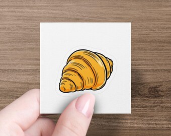 May include: A sticker depicting a cartoon croissant in golden-yellow with a black outline. The sticker is on a white square, held by a hand against a wooden background. The croissant has detailed lines.