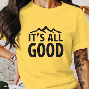 Può includere: Maglietta gialla con una grafica di montagna nera e il testo "IT'S ALL GOOD".