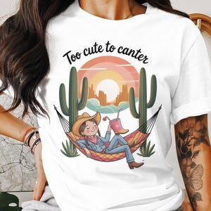 May include: White t-shirt with a desert scene graphic. The design shows a person in a cowboy hat relaxing in a hammock between cacti, with the text "Too cute to canter" above. The person is holding a drink.