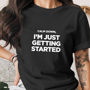 May include: Black t-shirt with the white text "CALM DOWN, I'M JUST GETTING STARTED". The shirt has short sleeves and a crew neck. The image shows a person wearing the shirt.