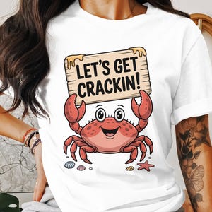 May include: White t-shirt with a cartoon crab holding a wooden sign saying "LET'S GET CRACKIN!". The crab is red and pink with a smiling face. Seashells and a starfish are at the bottom of the design.