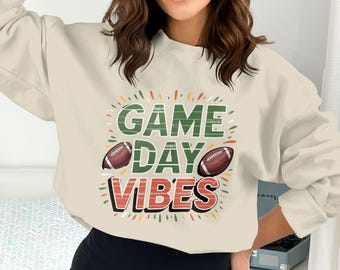 May include: A cream-coloured sweatshirt featuring the text "GAME DAY VIBES" in green, red, and orange, with two American footballs. The design is suitable for casual wear, ideal for game day.