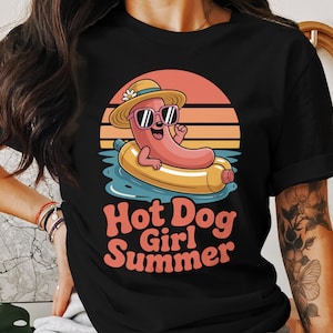 Pode incluir: Camiseta preta com um desenho animado de cachorro-quente usando óculos de sol e um chapéu de palha, flutuando em um inflável amarelo na água. O texto "Hot Dog Girl Summer" está abaixo, em um fundo de pôr do sol.