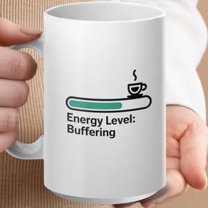 May include: White ceramic coffee mug with a black and teal graphic. The graphic depicts a coffee cup above a loading bar with the text "Energy Level: Buffering". The mug is held by a person.