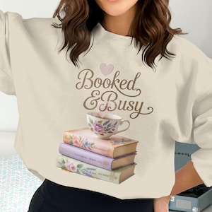 May include: Cream-colored sweatshirt with the text "Booked & Busy" and a heart graphic. A stack of books is topped with a teacup with floral designs. The books are in shades of pink and lavender.