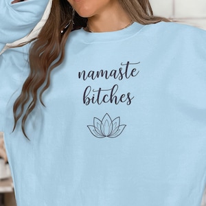Könnte beinhalten: Hellblaues Sweatshirt mit dem Schriftzug "namaste bitches" in Schreibschrift und einer Lotusblumen-Grafik. Das Sweatshirt hat eine einfarbige hellblaue Farbe.