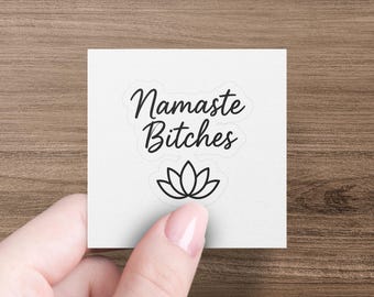 May include: A white square sticker with the words "Namaste Bitches" in a cursive font, above a lotus flower graphic. The sticker is held by a hand against a wooden background.