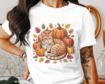 May include: White t-shirt featuring a watercolor illustration of a tabby cat curled up among pumpkins and autumn leaves. The pumpkins are various sizes and shades of orange and yellow, with green stems. The leaves are red, orange, and yellow.