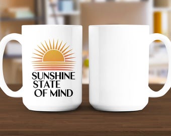 May include: White ceramic mug with a handle, featuring a graphic of a rising sun in shades of orange and yellow. The text "SUNSHINE STATE OF MIND" is printed below the sun graphic in black. The mug is displayed on a wooden surface.