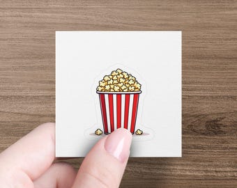 May include: A sticker featuring a cartoon illustration of a popcorn bucket. The bucket is red with white vertical stripes and filled with fluffy, yellow popcorn. Several popcorn kernels are scattered around the base of the bucket.