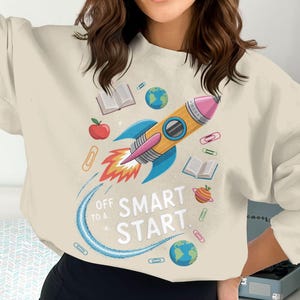 Może przedstawiać: Kremowa bluza z kolorową grafiką przedstawiającą rakietę, książki, jabłka i globusy. Tekst "Off to a Smart Start" jest wydrukowany poniżej. Projekt jest zabawny i edukacyjny.
