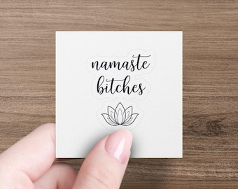 May include: A white square sticker with the words "namaste bitches" in black script above a black line drawing of a lotus flower. The sticker is held by a hand against a wood grain background.