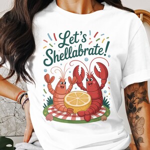 Könnte beinhalten: Weißes T-Shirt mit einem Cartoon-Design von zwei lächelnden Hummern und einer Zitronenscheibe. Der Text "Let's Shellabrate!" steht über den Hummern. Bunte, konfettiartige Akzente umgeben das Design.