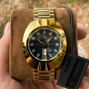 May include: A gold-toned Rado Diastar watch with a black face and diamond hour markers. The watch has a gold band and a date display. A black tag with the Rado logo is attached.