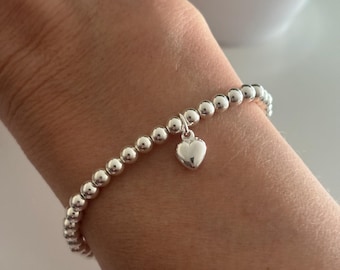 Peut inclure: Un bracelet composé de perles argentées uniformes et réfléchissantes, agrémenté d'une petite breloque en forme de cœur lisse et polie suspendue au centre. Les perles sont enfilées pour former une boucle circulaire continue.