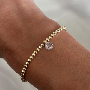 May include: A stretch bracelet composed of small, polished gold-toned spherical beads arranged in a continuous circular pattern, featuring a single, clear, faceted circular crystal charm suspended from the centre of the bead strand.