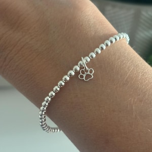 May include: Silver beaded bracelet with a paw print charm. The bracelet features small, round silver beads strung together, with a delicate paw print charm hanging from the centre. The bracelet is designed to be worn on the wrist.
