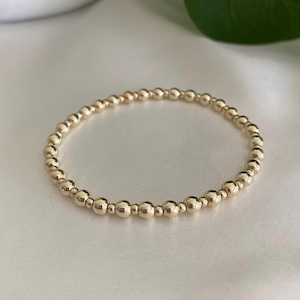 May include: A gold beaded bracelet. The bracelet features small, round, gold-coloured beads strung together. The beads have a shiny finish and are uniform in size. The bracelet is displayed on a white, silky surface.