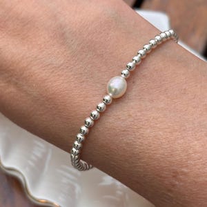 May include: A stretch bracelet featuring a single white oval freshwater pearl centred among numerous small, round, polished silver-toned metallic beads. The beads are arranged in a continuous circular pattern, creating a simple and uniform design.