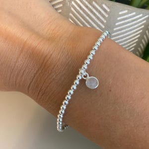 May include: A stretch bracelet composed of small, reflective silver-toned spherical beads. The bracelet features a single, circular charm with a smooth, translucent, pale iridescent stone set in a silver-toned metal frame, dangling from the band.