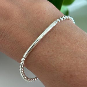 May include: A silver bracelet with a bar and bead design. The bracelet features a central, rectangular bar and is accented with small, round beads. The jewellery is worn on a wrist.