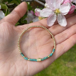 May include: A gold and turquoise beaded bracelet with a gold charm. The bracelet is on a person's wrist.