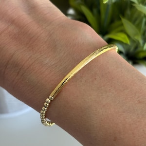 May include: A gold bracelet featuring a textured bar and a beaded section. The bracelet is displayed on a wrist, highlighting its simple design. The background includes green foliage, adding a natural touch.