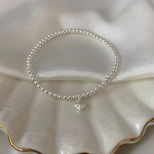 May include: A silver beaded bracelet with a small heart charm. The bracelet is made of small, round beads and is displayed on a white, textured surface.