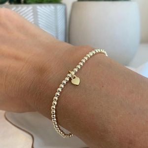 May include: A stretch bracelet composed of small, polished gold-tone spherical beads arranged in a continuous circular pattern. A single, smooth, flat gold-tone heart-shaped charm hangs from the centre of the beaded strand.